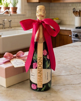 A Chandon sparkling wine bottle decorated with a large bright pink ribbon bow, standing on a marble kitchen counter next to a pink and cream gift box with a matching bow. The kitchen background features wooden cabinets, a white farmhouse sink, and potted plants.